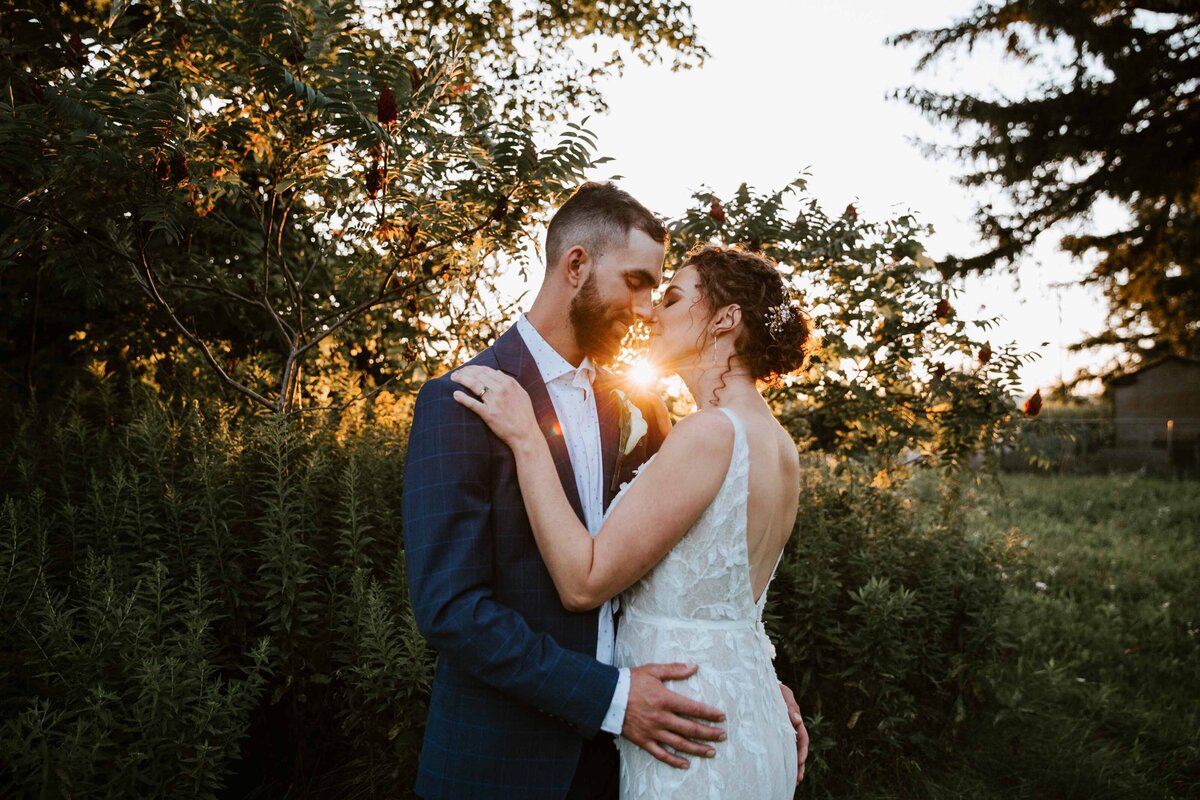 Bride and groom kissing at sunset. The sun is peek between them. there is a rustic barn in the distance.