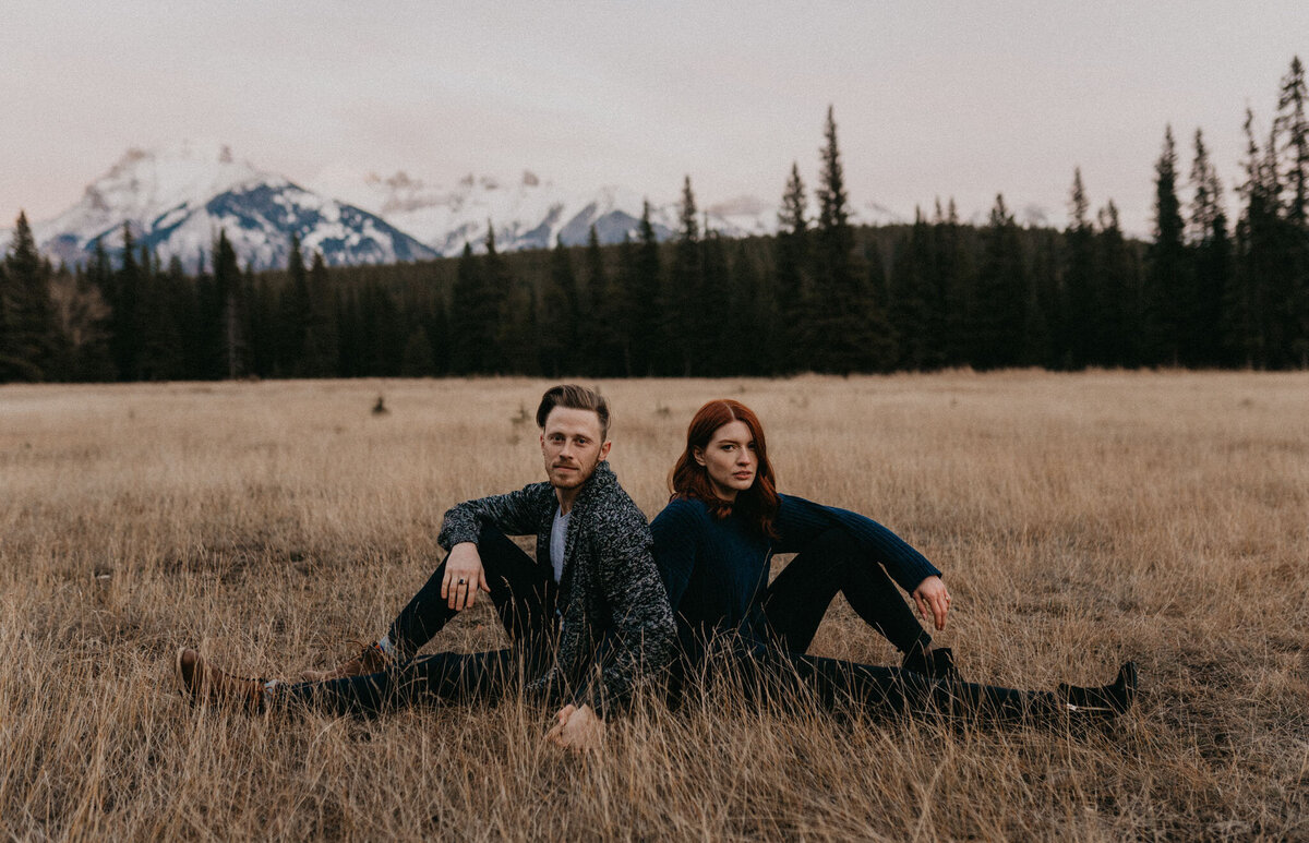 Couple sitting back to back in a field in the mountains of Alberta, captured by Ash Maclean Photography, romantic elopement and wedding photographer in Red Deer, Alberta. Featured on the Bronte Bride Vendor Guide.
