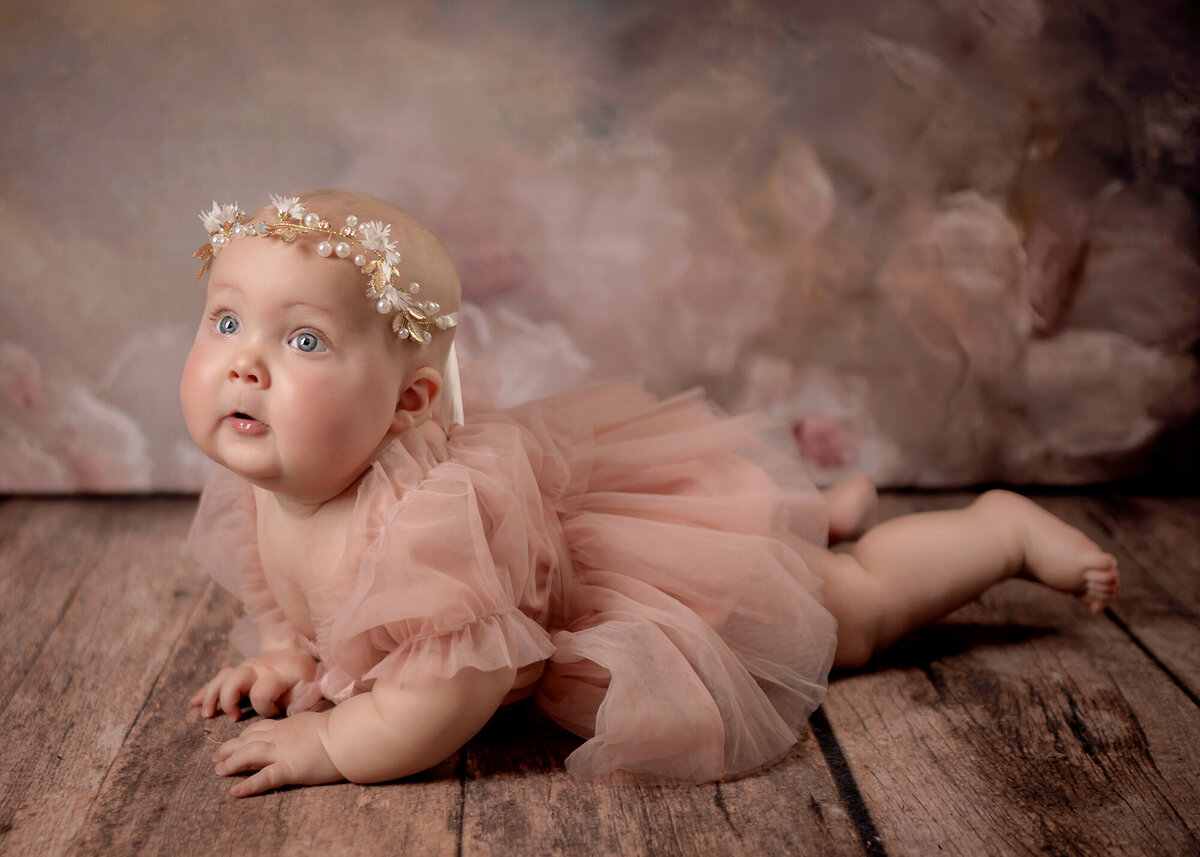 Laying on a wooden floor, a baby looks away in her pink dress