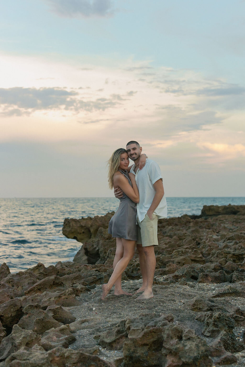 A south florida couple embraces at a Jupiter beach with rocky shoreline during blue hour.