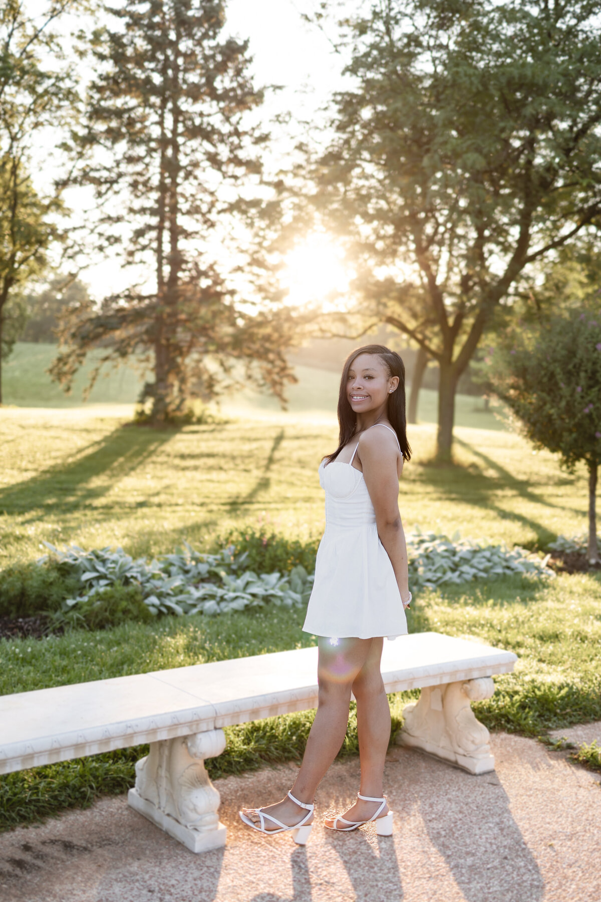 a young woman wears a white dress and stands facing away from the camera with her hands behind her back. The bright green grass and sun rays are in the backgroud 