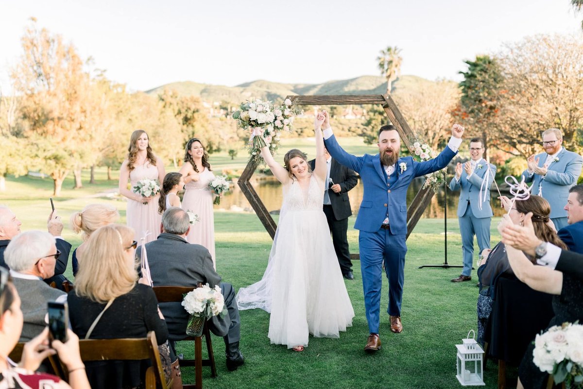 Bride and Groom walk down the aisle between guests with arms in the air in celebration of their marriage