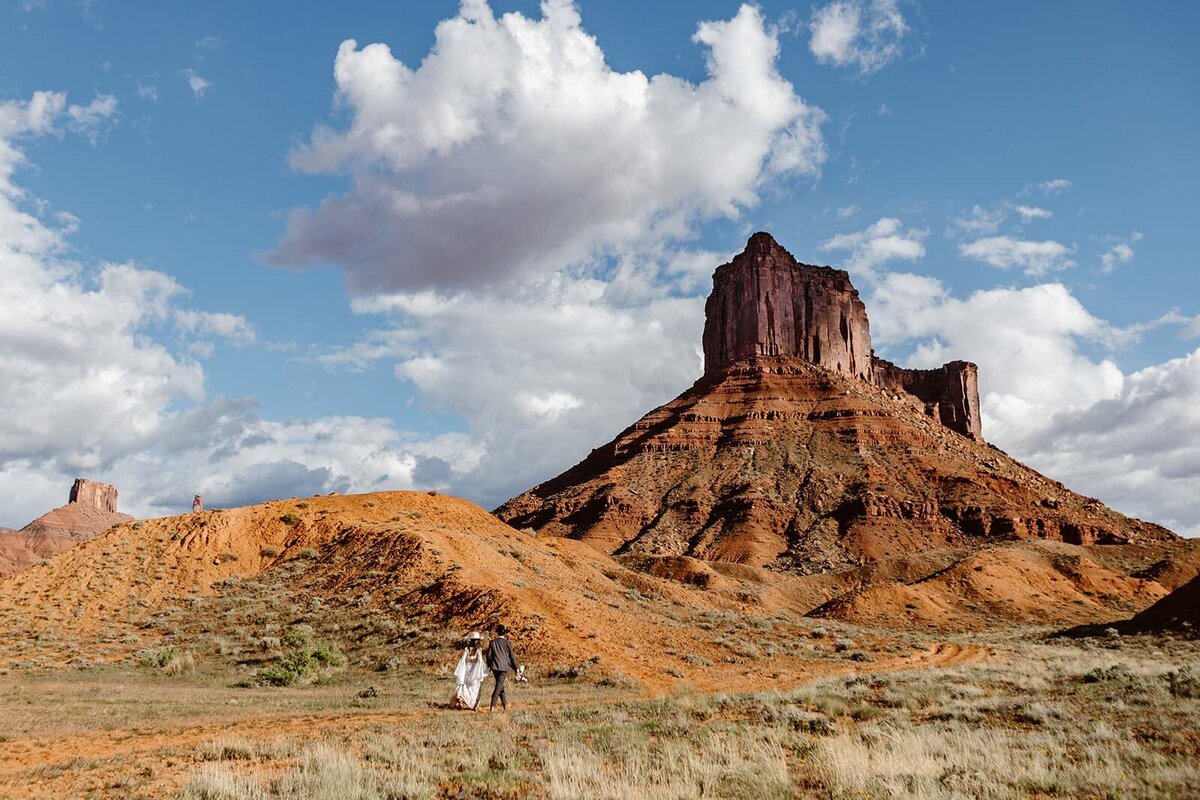 A bride and groom walk up a trail at Ida Gulch in Moab. They are walking towards a towering red rock structure. 