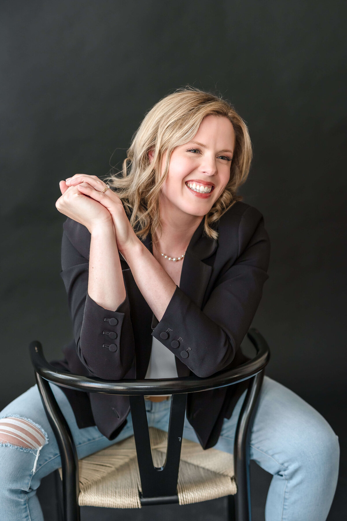 Laughing female entrepreneur leaning on black chair wearing black blazer and ripped jeans in Kelowna photo studio.