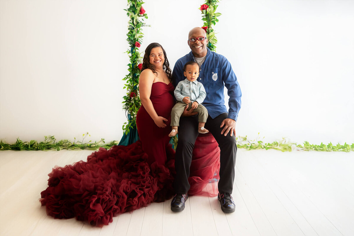 A man and pregnant woman in a red dress happily pose with their adorable baby.