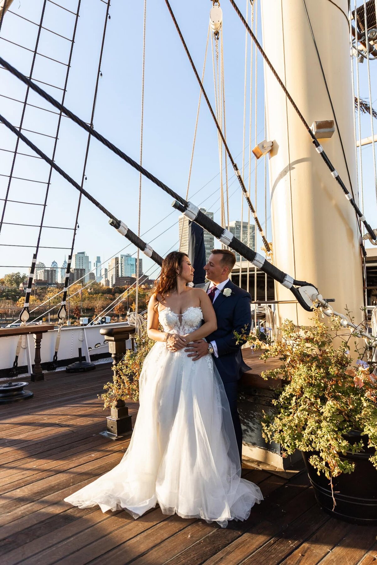 The bride and groom stand on the deck of The Moshulu, a historic tall ship docked along the Philadelphia waterfront, with the city skyline rising behind them. The bride’s flowing wedding dress catches the breeze as the groom, dressed in a sharp navy suit with a white boutonniere, wraps his arms around her, savoring a quiet moment on their wedding day