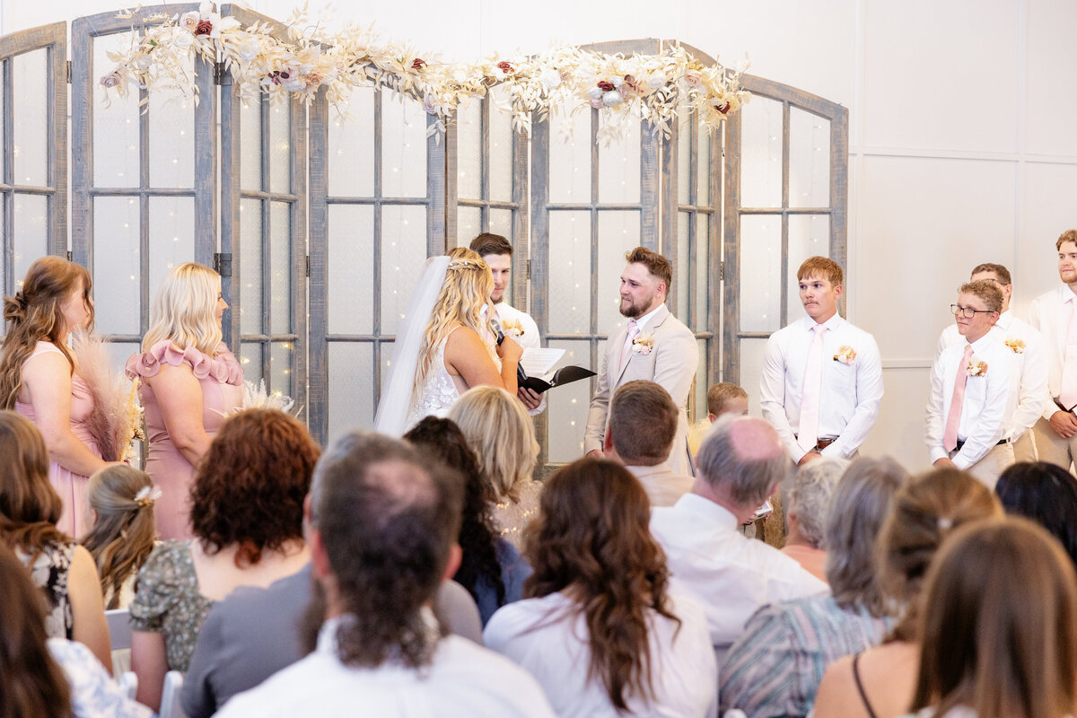 The bride, holding a microphone, reads her heartfelt vows to her groom, who listens with a loving smile. The wedding party and guests witness this emotional moment under a beautifully adorned rustic wooden arch.