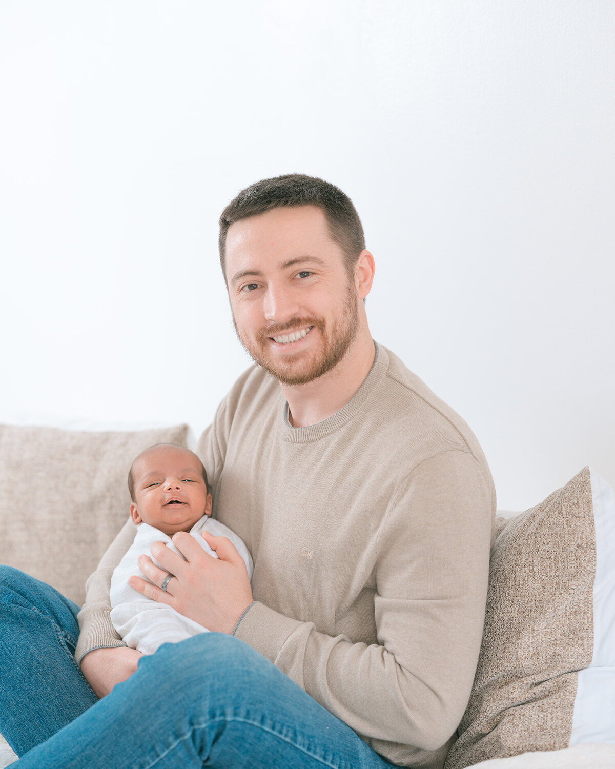 Father wearing light brown sweater sitting on white bed holding newborn baby and smiling at camera, captured in natural light by Menlo Park newborn photographer during in-home session.