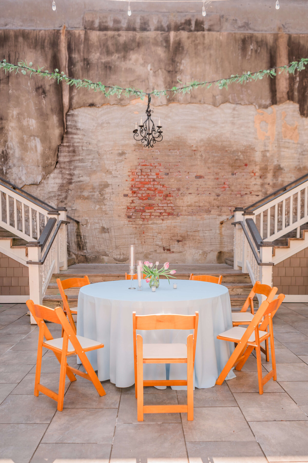 A round table with a light blue tablecloth, a floral centerpiece, and six wooden chairs is set outdoors on a stone patio, with greenery and a white railing in the background. 