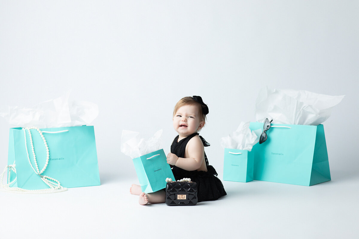 Dallas milestone photographer captures a stylish baby girl in a black lace outfit sitting among Tiffany blue gift bags, holding one bag with a bright smile and a tiny black purse at her feet.