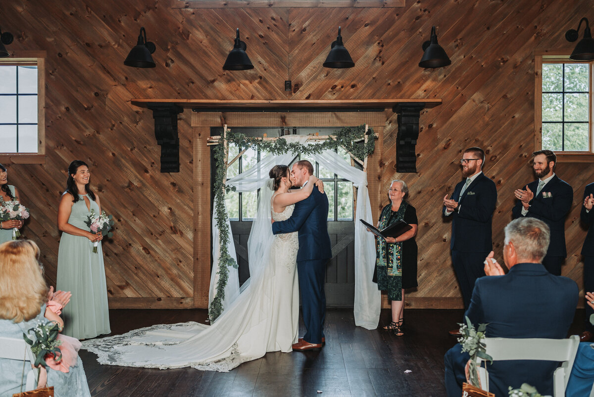 Bride and groom share their first kiss under a floral arbor during an indoor ceremony at Sweeney Barn in Manassas, Virginia.