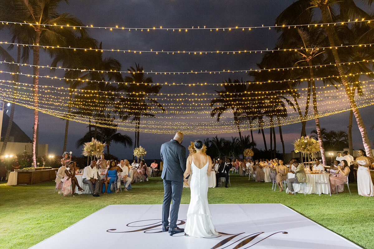Bride and groom stand hand in hand on a custom dance floor under glowing string lights at their destination wedding in Puerto Vallarta, Mexico. The tropical outdoor reception features elegant decor, palm trees, and a sunset sky, creating a romantic and luxurious wedding atmosphere.