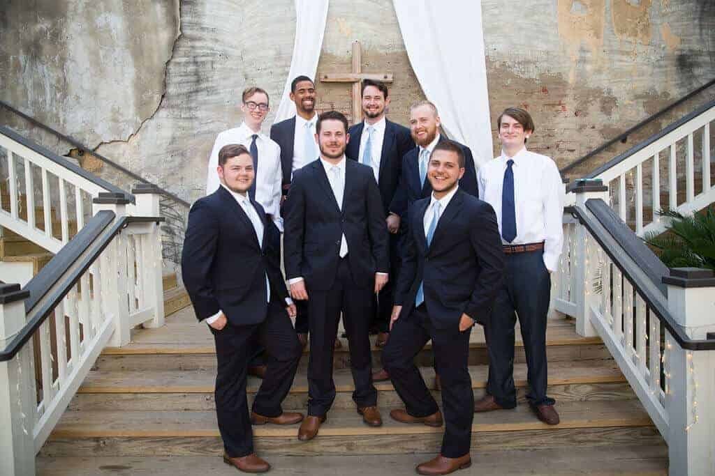 A group of eight men in dress shirts, ties, and suits pose together on outdoor wooden stairs with white railings, in front of a brick wall draped with white fabric and a wooden cross.