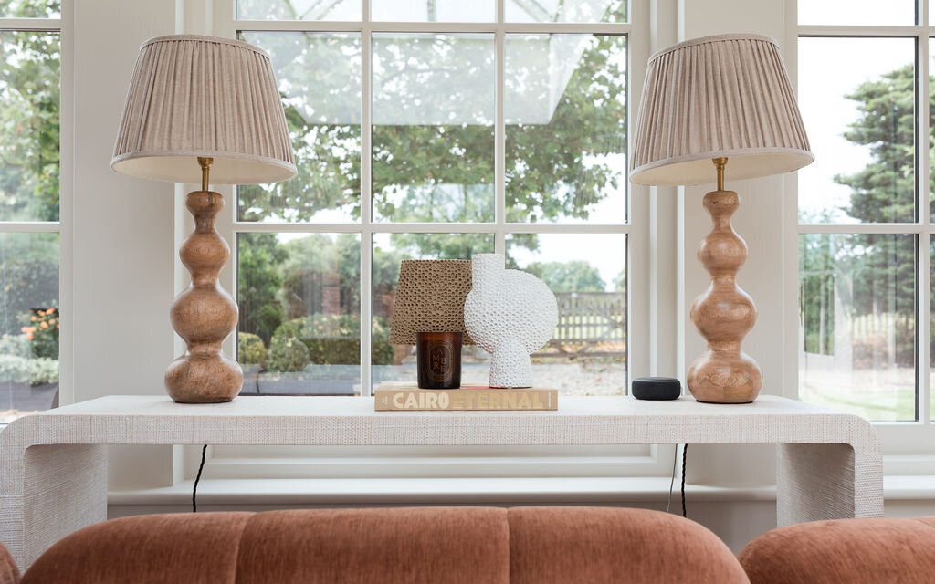 A console table nestled behind a plush, rust orange sofa. Two large neutral lamps sit on the table. Behind the table are lage windows, showing greenery outside.