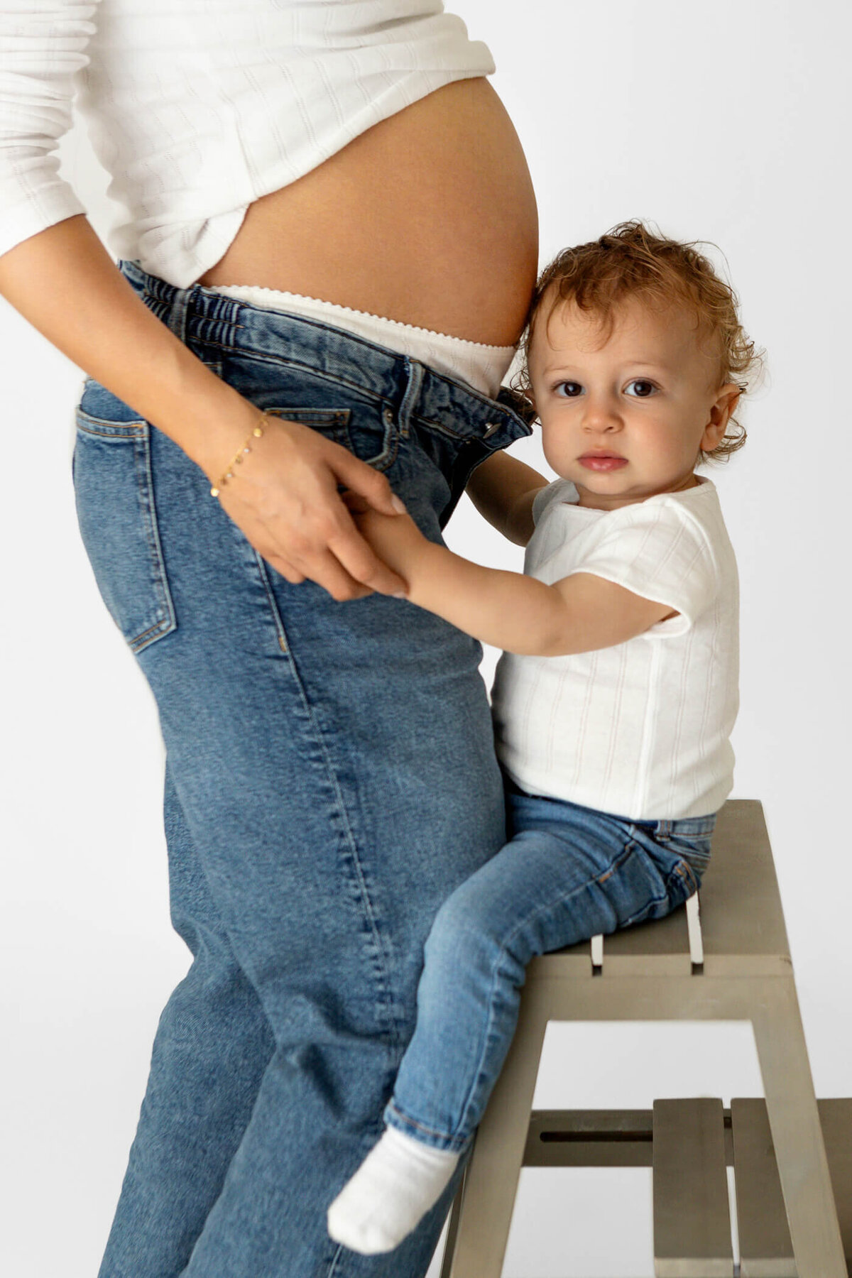 A toddler with curly hair sits on a stool, holding onto the waistband of a pregnant woman's jeans. The woman is standing next to the child, wearing a white top that reveals her bare baby bump above the jeans. The toddler, dressed in a white shirt and blue jeans, looks directly at the camera with a calm expression, while the woman's face is not visible in the frame. The image captures a tender moment between mother and child, emphasizing the connection and anticipation of the new addition to the family.