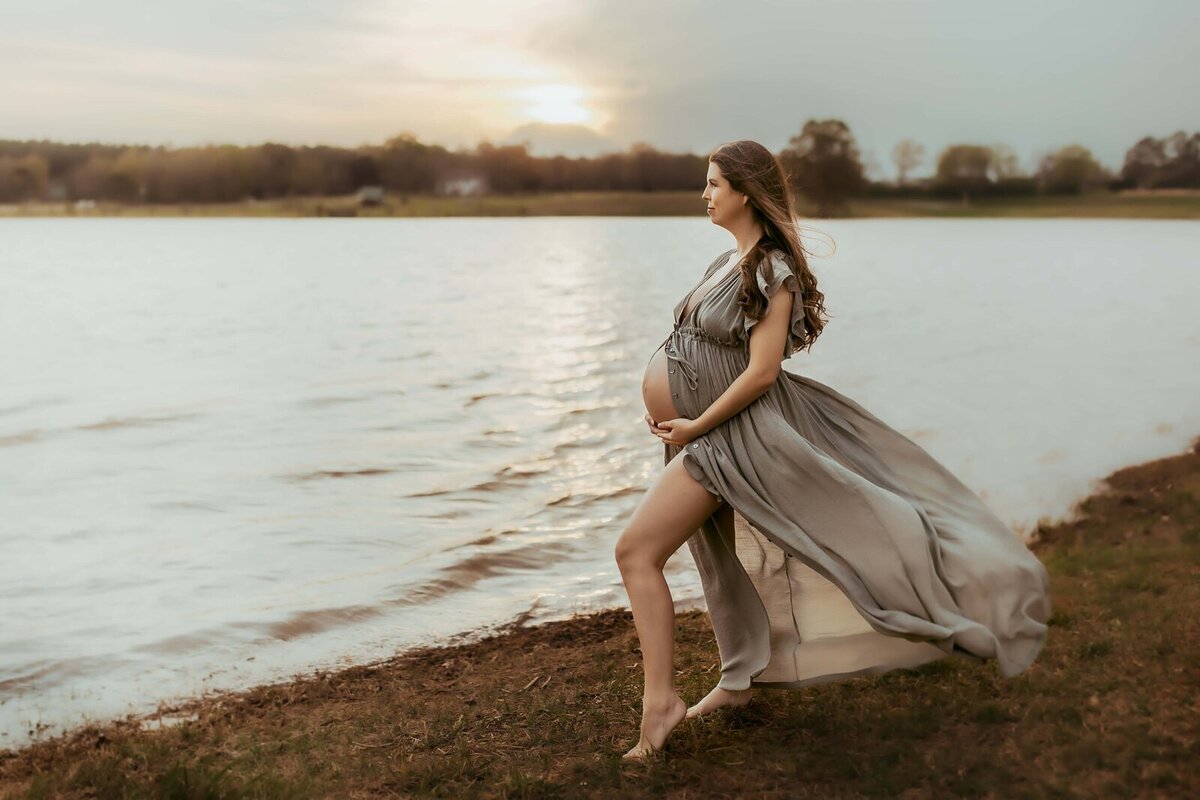 Pregnant woman in a flowing dress stands barefoot by a lake at sunset, looking into the distance.
