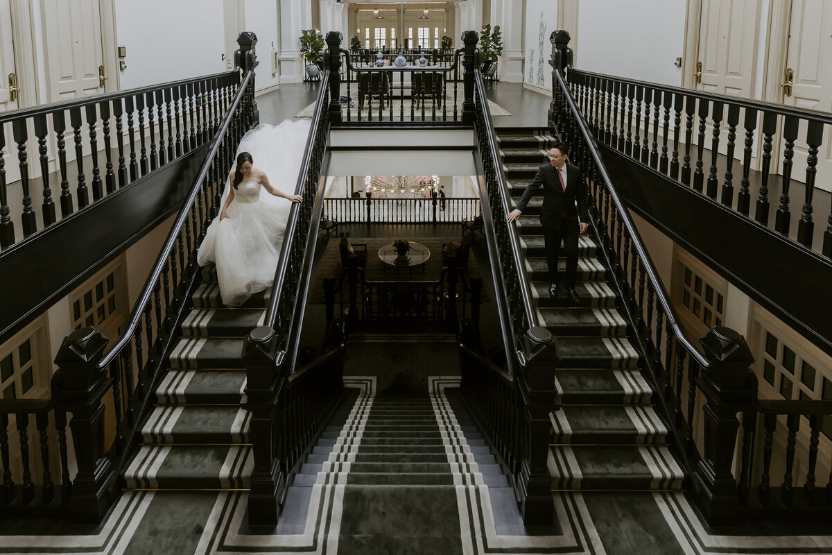 bride in a wedding dress with a long train and groom wearing a black suit walk down the stairs of the raffles hotel