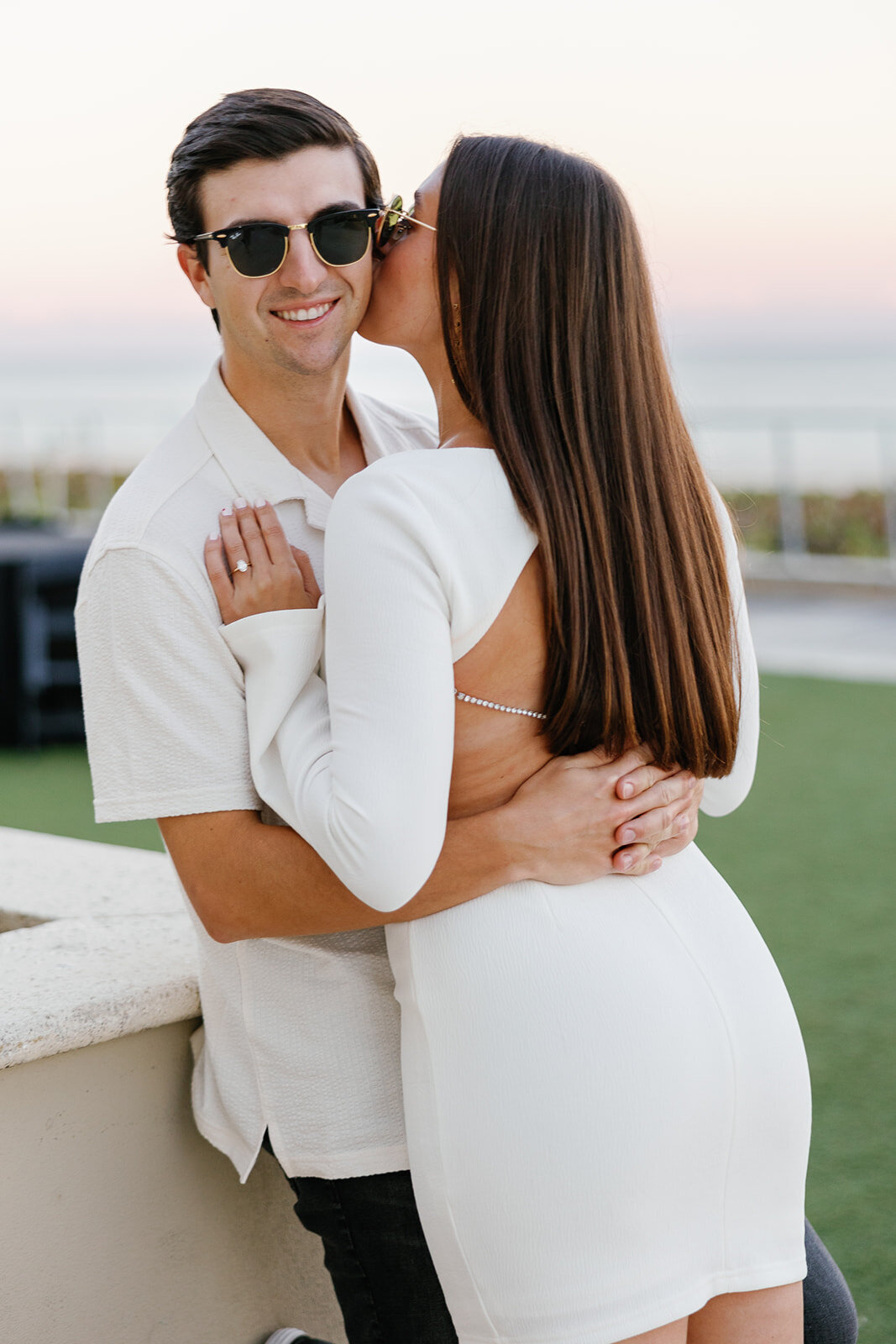 Rebecca, wearing an elegant white dress, kisses Graysen on the cheek while he smiles confidently at the camera, showcasing their love during their sunset engagement session at Marriott Hotel Fort Lauderdale.
