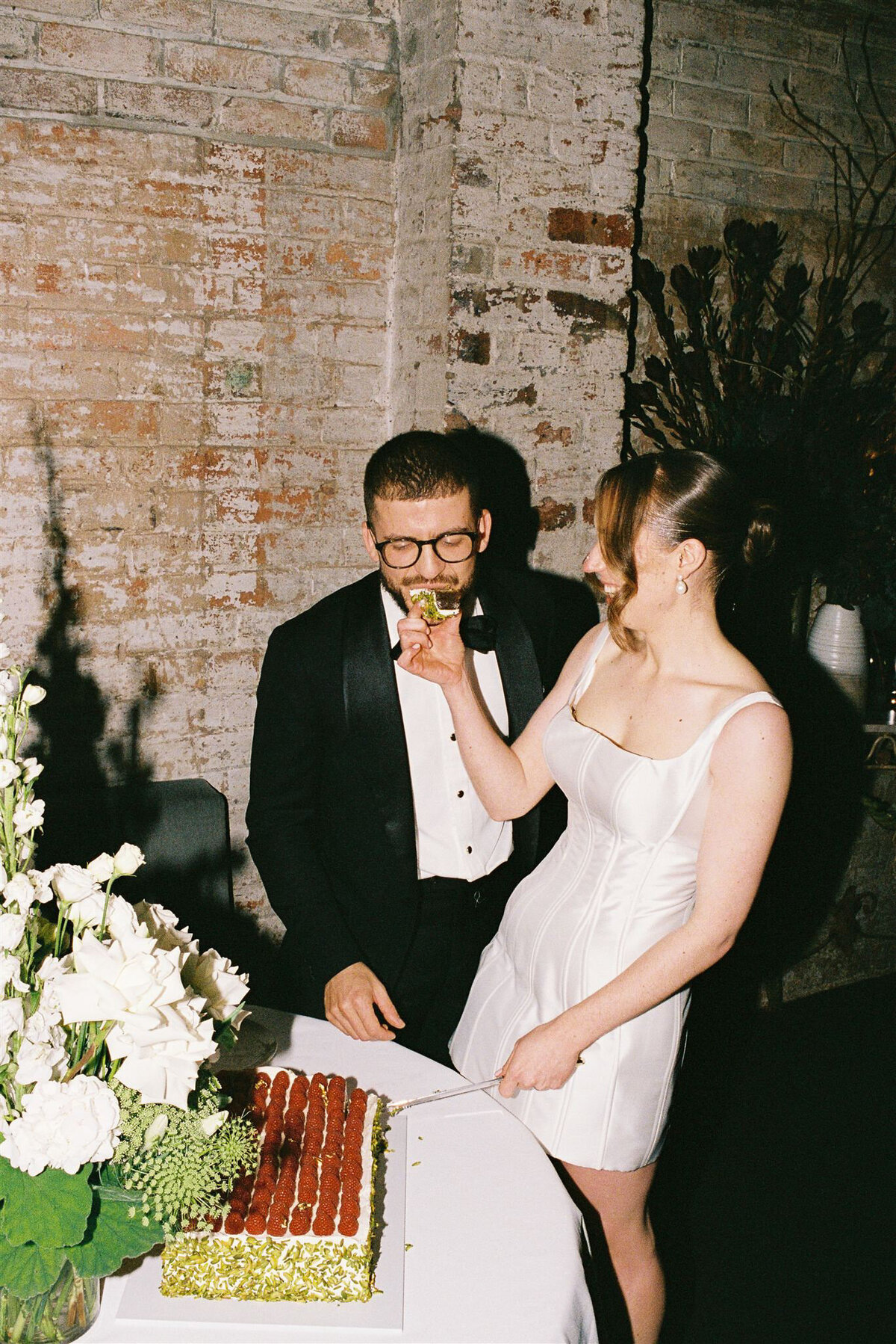 Newlyweds cutting their wedding cake and feeding each other, captured candidly on 35mm film at their intimate celebration.