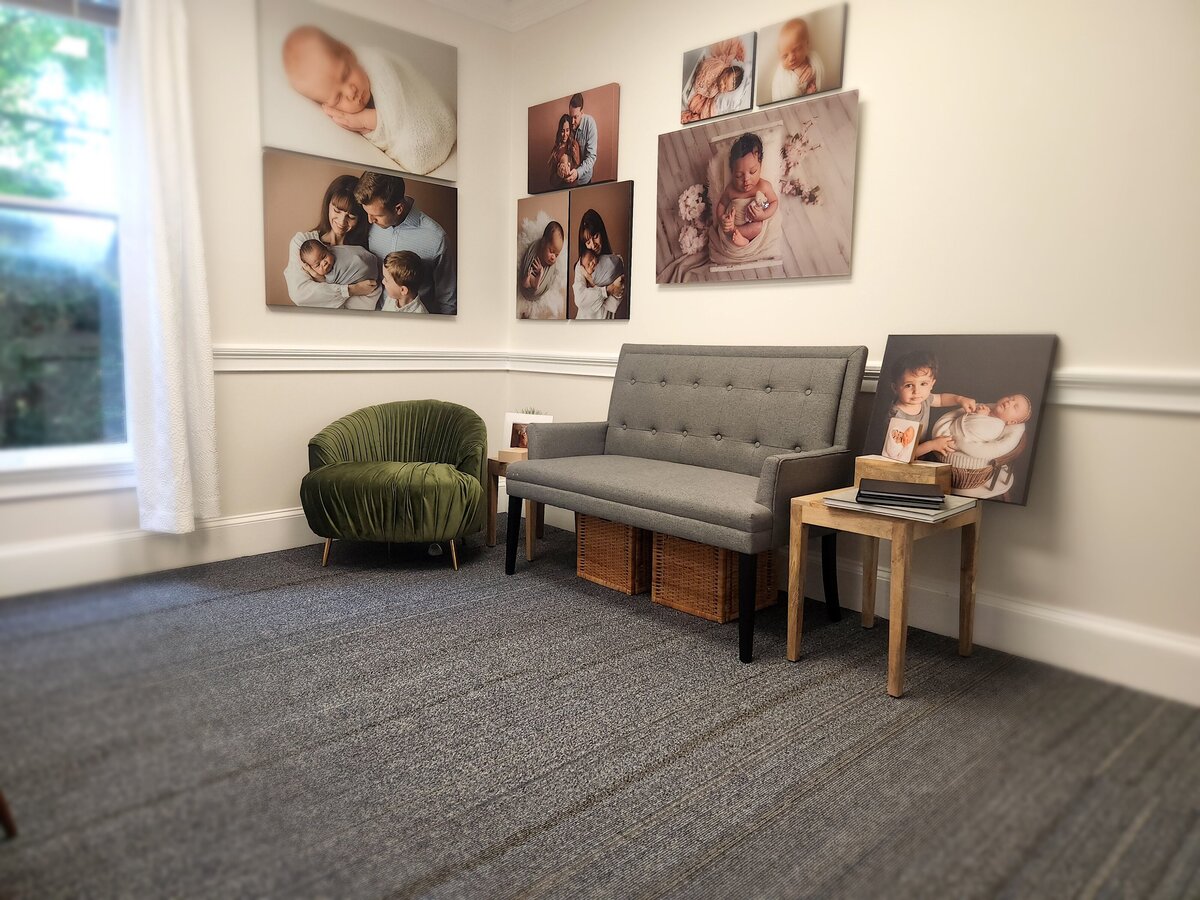 Office and ordering appointment area with white walls, olive green mid century modern upholstered chair, gray setae, wooden end tables and large pieces of wall art of babies covering the walls