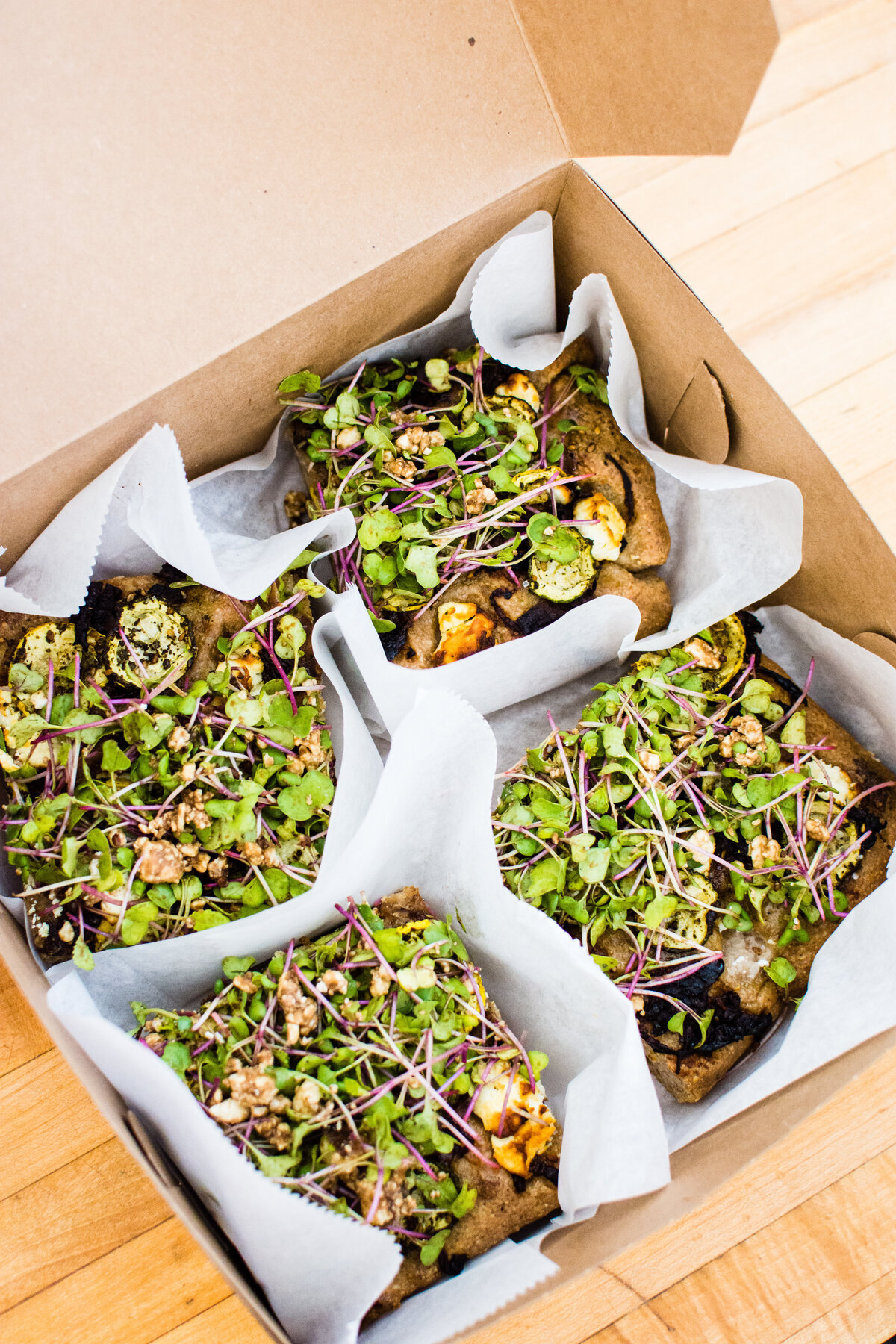 Focaccia with savory toppings and micro greens tucked in parchment paper in a brown box