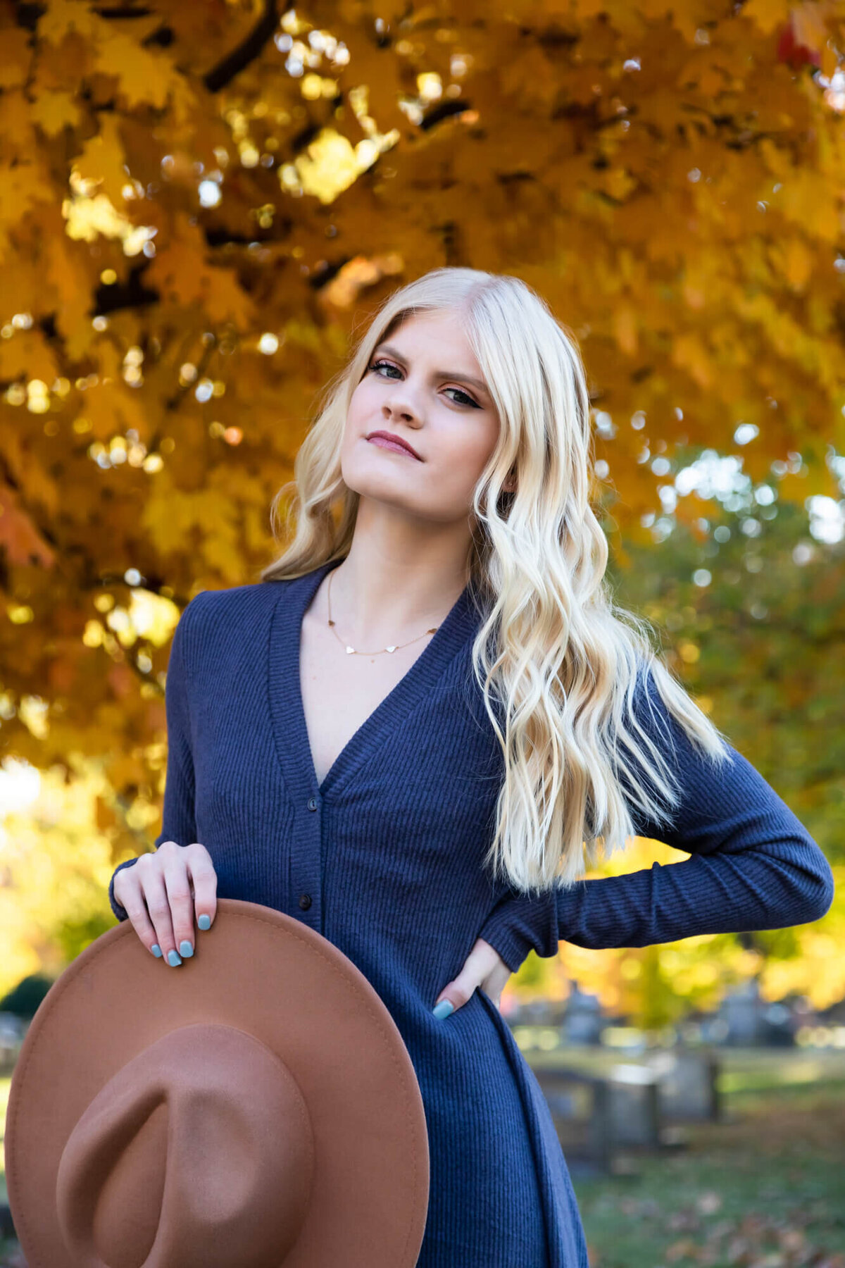A lovely senior portrait of a beautiful blonde girl wearing a blue dress and posing with a brown hat in front of golden Fall leaves. Captured by Springfield, MO senior photographer Dynae Levingston.