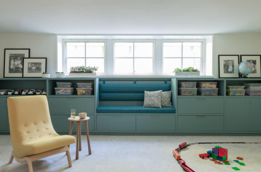 Boston decorator designs a green built-in playroom with yellow lounge chairs, open space for play, shelves for toys, a custom bench, and Brook Perdigon fabric cushions.
