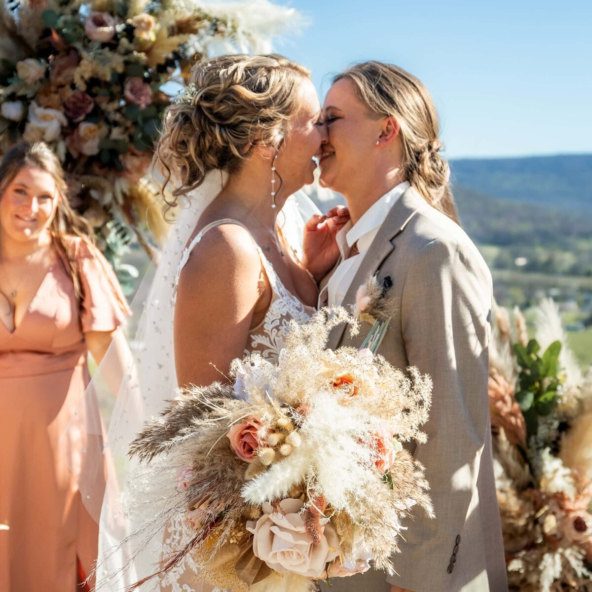 Brides kiss on wedding day while family and friends look on and smile, this lesbian wedding took place overlooking a gorgeous mountain view with large floral arrangements.