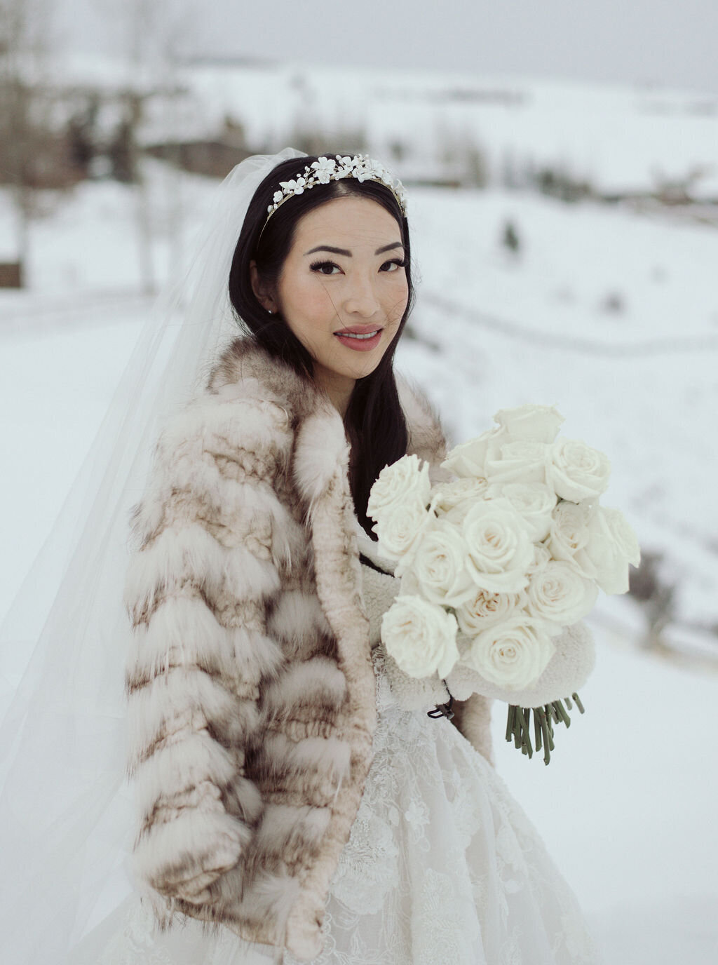 Jackson Hole winter bride smiles with a bouquet of white roses and a warm fur on before meeting her husband for their elopement below the Grand Tetons in Wyoming