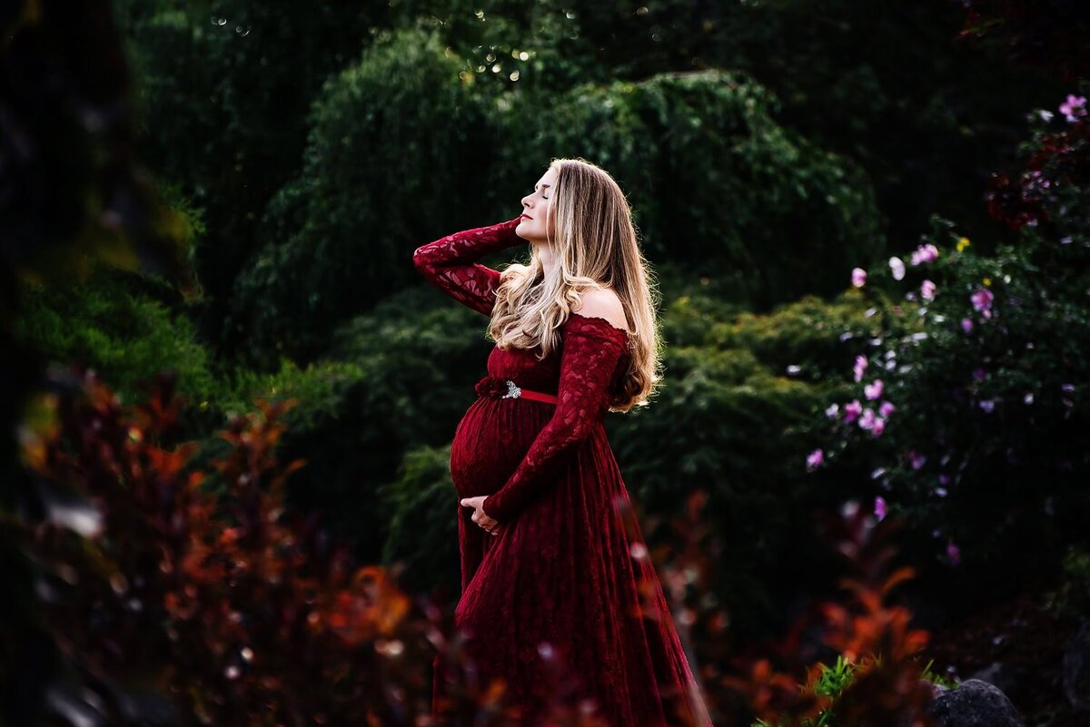 Expecting mother in a red lace dress striking a dramatic pose during an outdoor maternity session in the garden at Deer Lake Park.