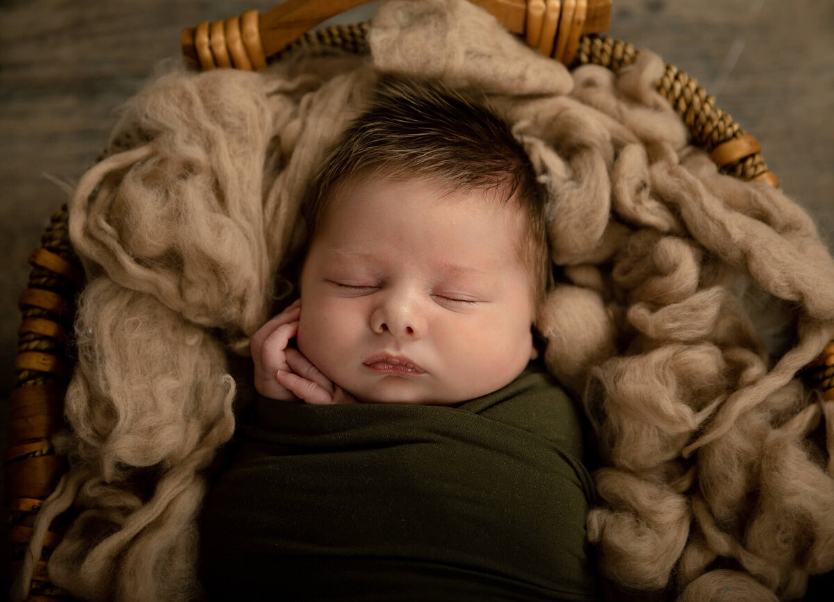 Newborn boy with lots of light brown hair laying in a basket with light brown wool surrounding his.