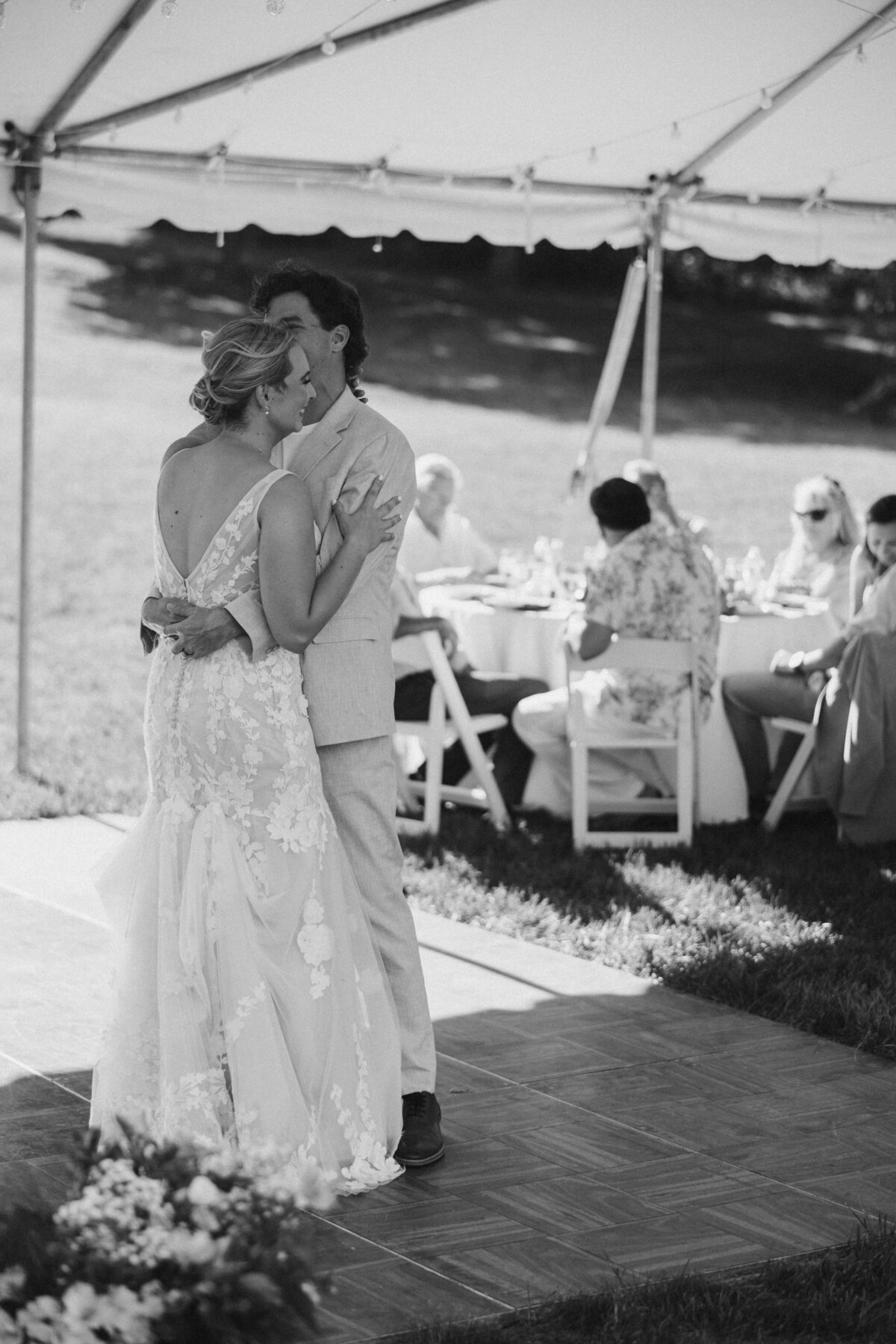 Bride and groom share a romantic first dance under a tent at their outdoor wedding, with guests seated at round tables in the background.