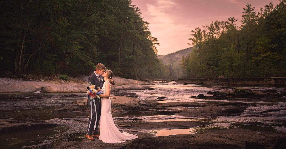 A wedding couple kissing as they stand on rocks next to a river.