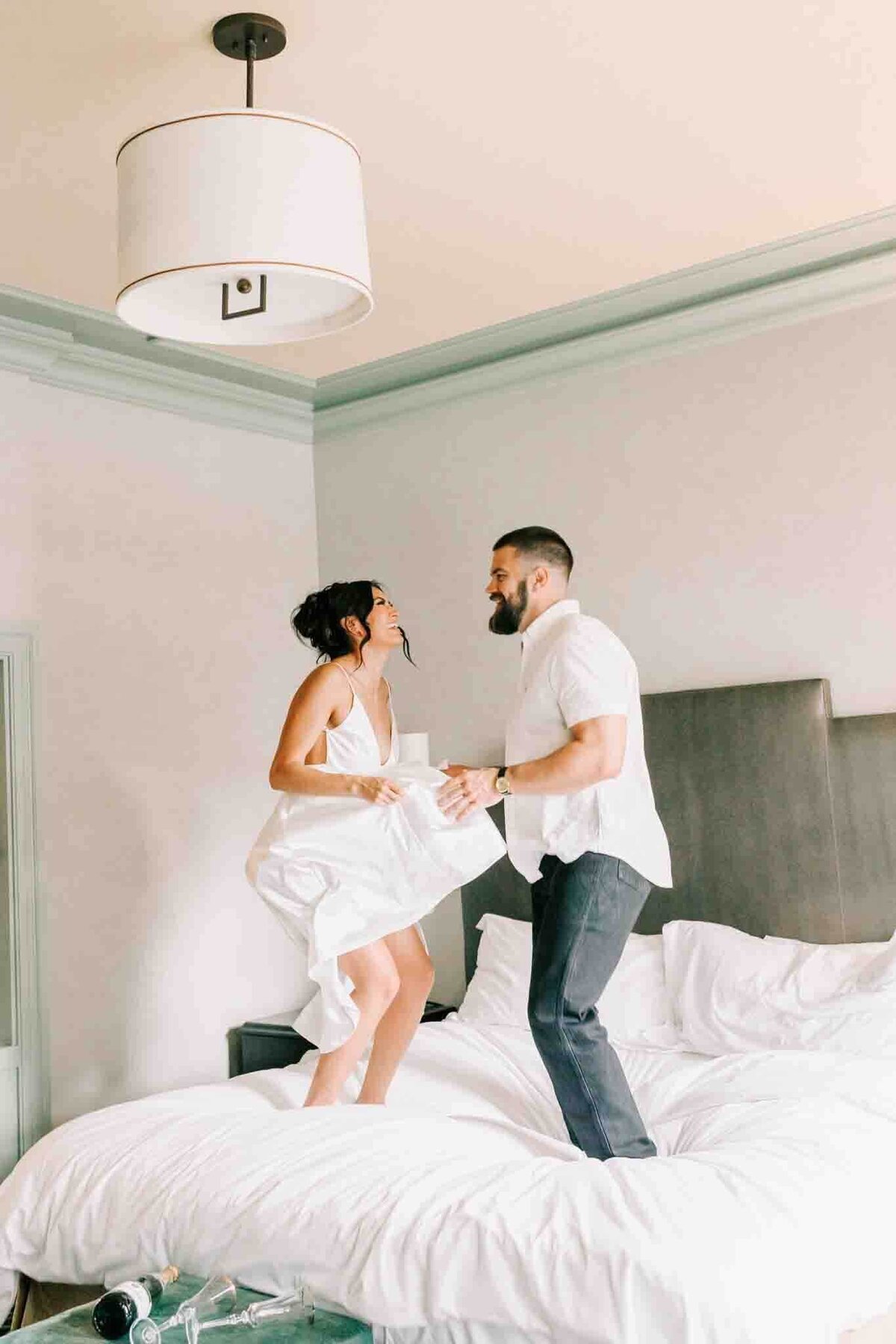 bride and groom celebrate their engagement by jumping on top of a bed in a fun hotel room