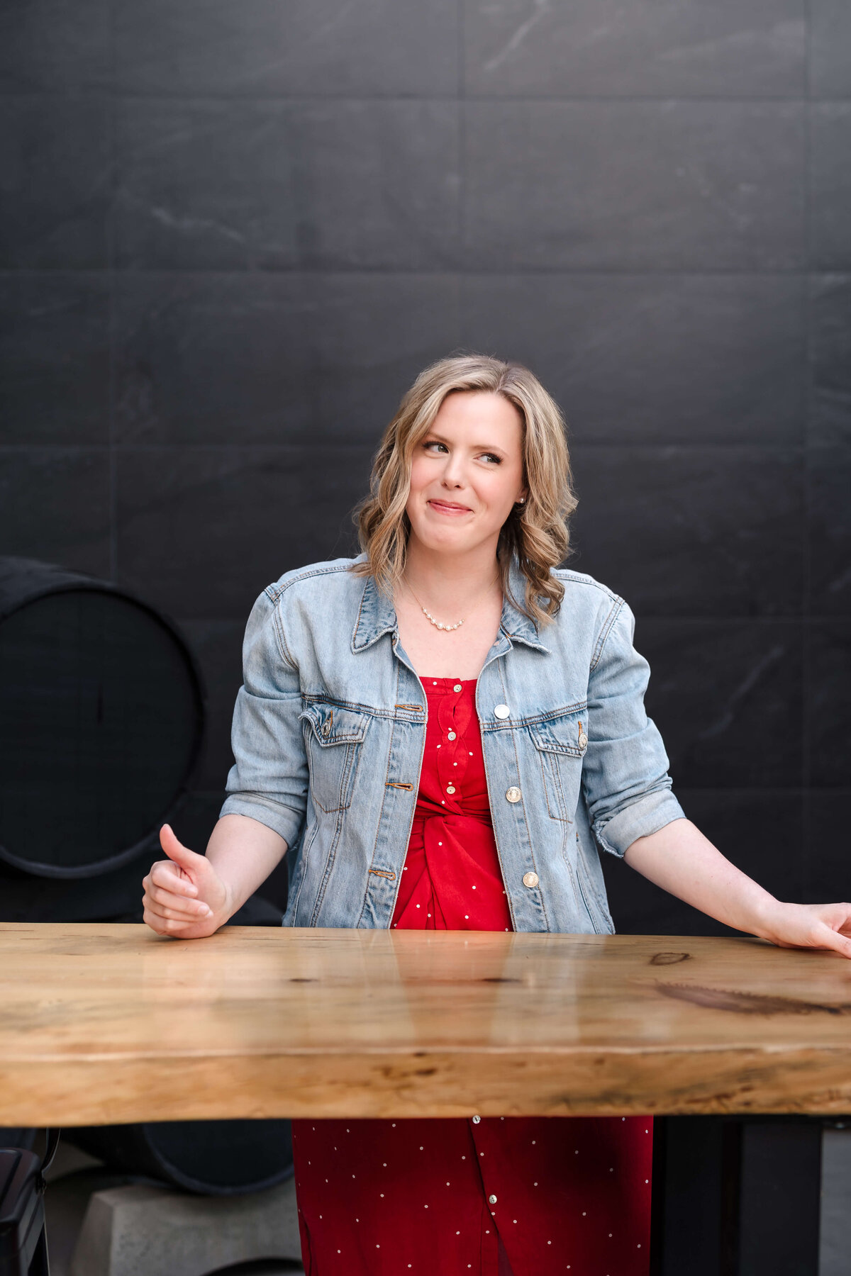 Skeptical female entrepreneur standing behind wooden table wearing denim jacket over red dress in Kelowna.