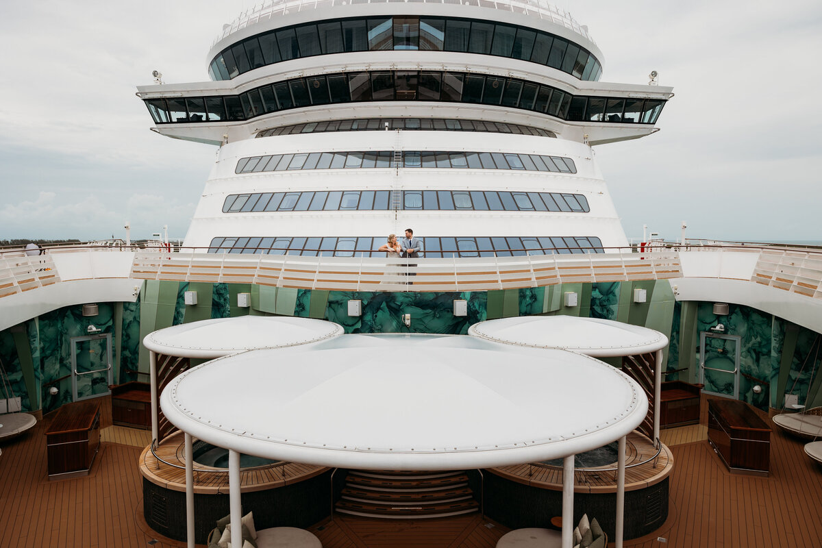 Bride and groom stand on an upper deck of the Disney Wish, nestled between sweeping curves of the ship’s architecture with hidden mickey detail