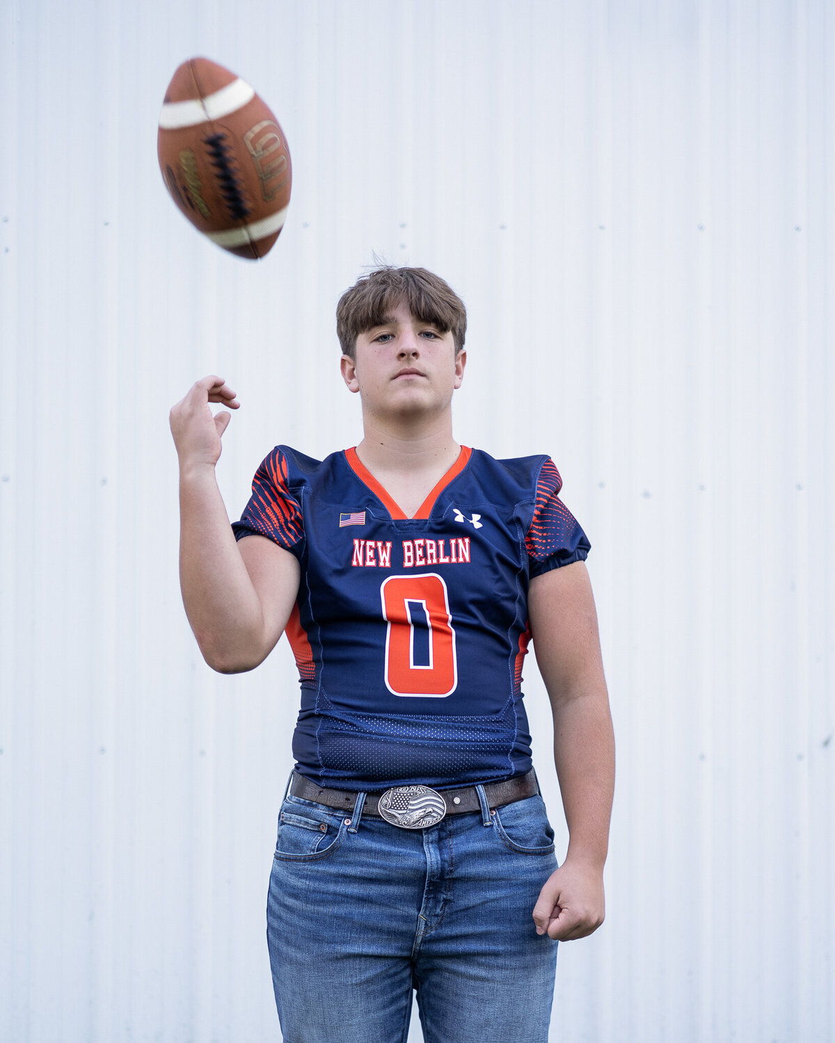 High school senior male in a navy and orange football jersey, blue jeans, belt, looking at the camera with a serious look, tossing up a football and has short brown hair