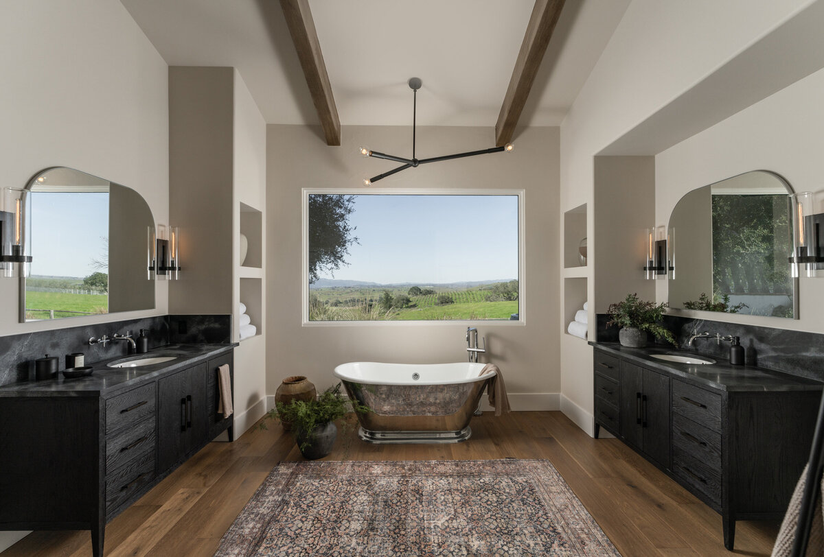 primary bathroom with view of a vineyard free standing tub silver tub counter balance light wood ceiling beams black vanity dark hardware soapstone countertop arched mirror vintage rug vintage vases integrated built-in