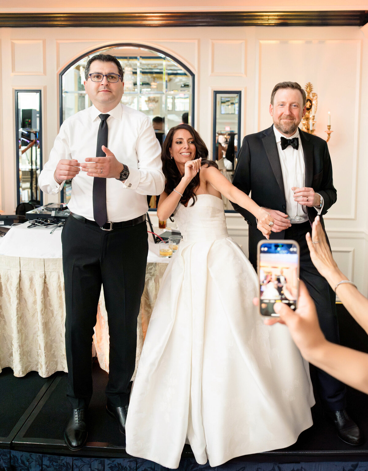 A wedding scene with three people: a bride in a white dress playfully pulling the tie of a groom in a tuxedo, while another man in a white shirt watches. A hand in the foreground holds a smartphone, capturing the moment.