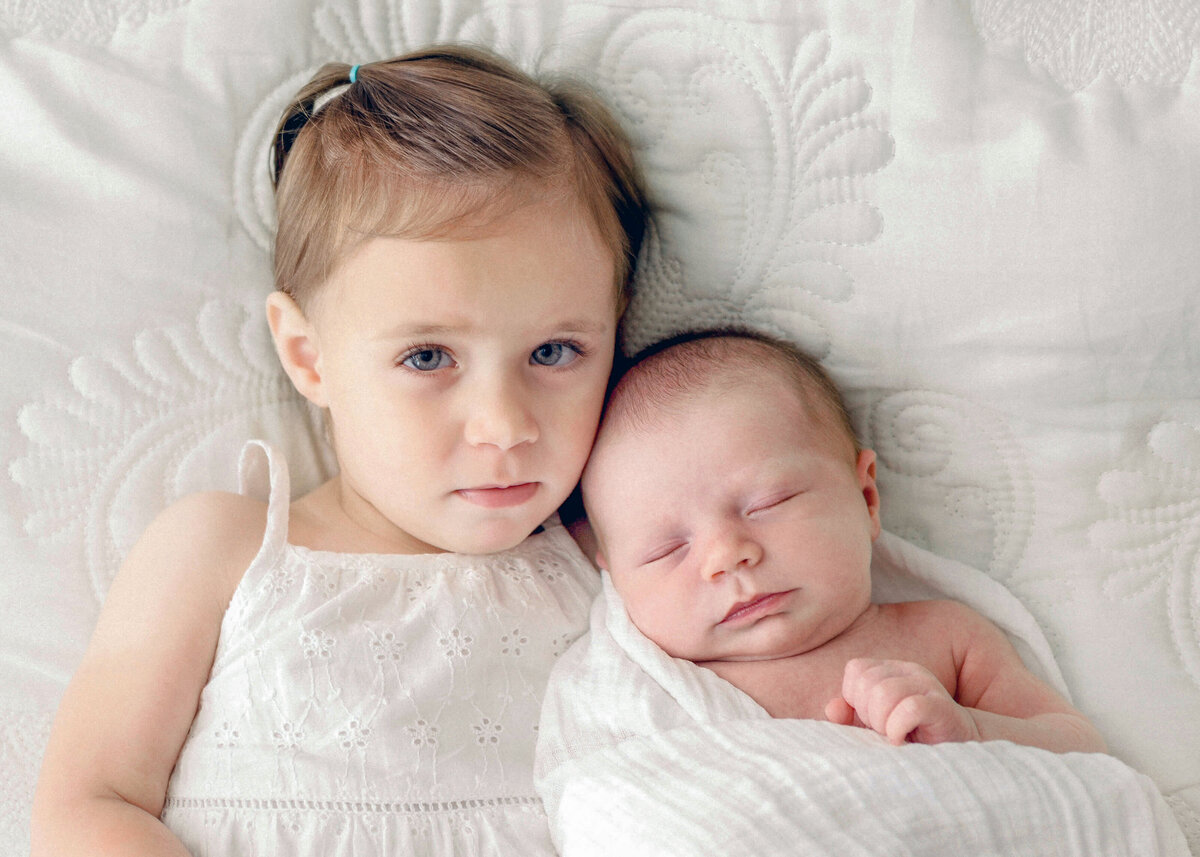 A shy toddler girl in a white dress holding newborn baby in white blanket on white comforter.