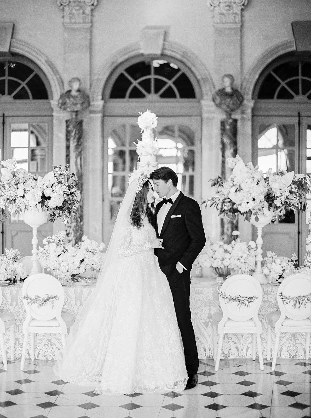" Here is a captivating black-and-white photo captured at the enchanting Chateau Vaux le Vicomte in Paris, France. Renowned photographer Audrey FORA has beautifully framed this moment at one of the top destination wedding venues in France. The image features a bride and groom standing closely together, gently touching foreheads. The bride looks stunning in a long lace gown and veil, contrasted by the groom's classic black tuxedo. They are enveloped by exquisite ornate floral arrangements, creating an elegant and romantic ambiance. In addition, East West Films is also on-site as the videographers, ensuring every special moment is captured on film."