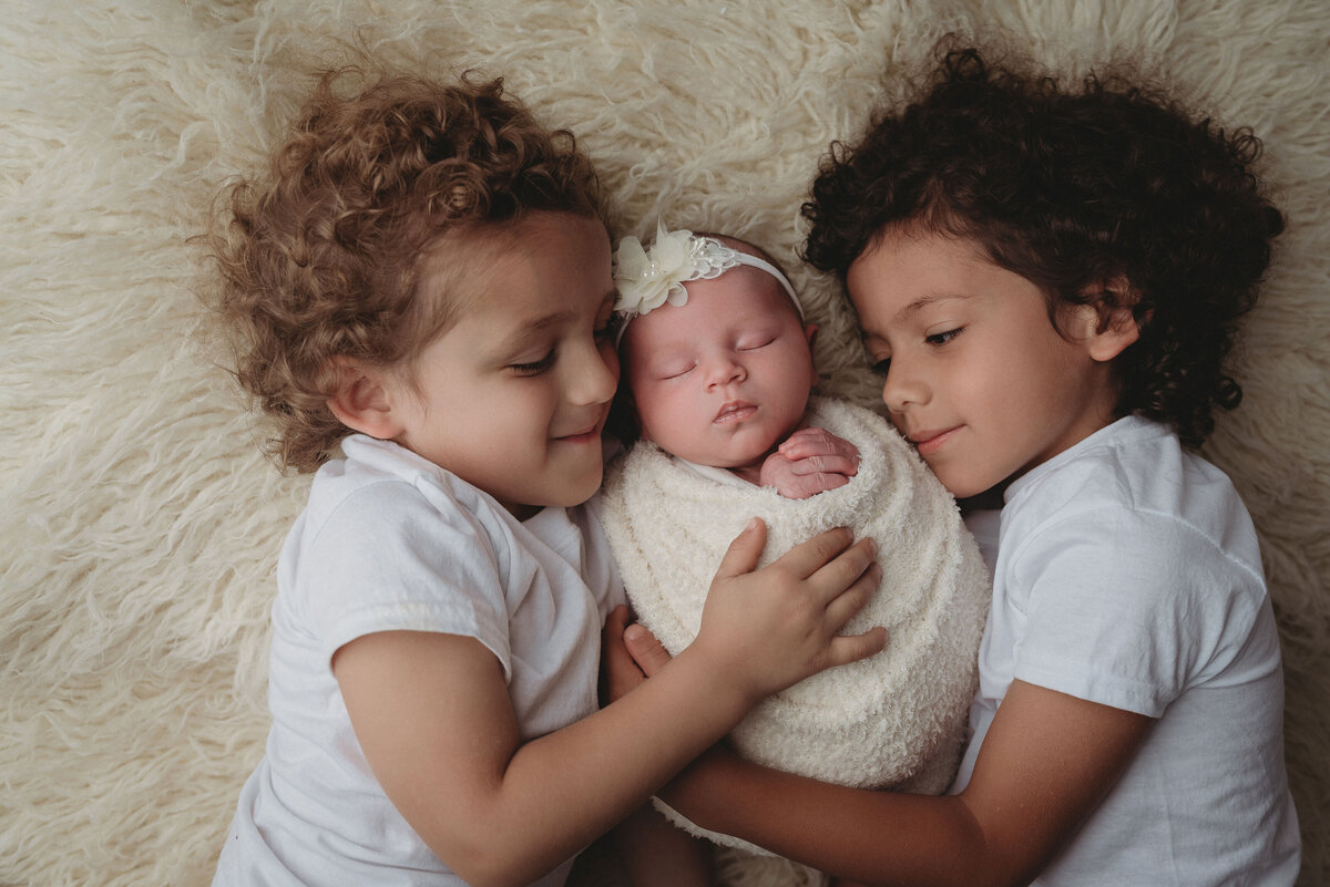 Newborn baby girl swaddled in white sleeping on back laying in between big brothers who are hugging baby.