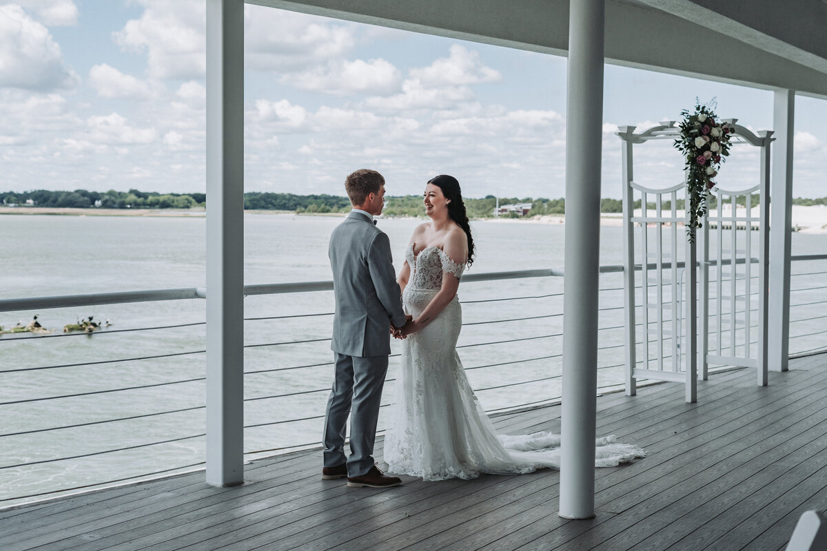 Bride and groom hold hands under a floral arbor on the balcony of Lesner Inn with a waterfront view in Virginia Beach, Virginia.