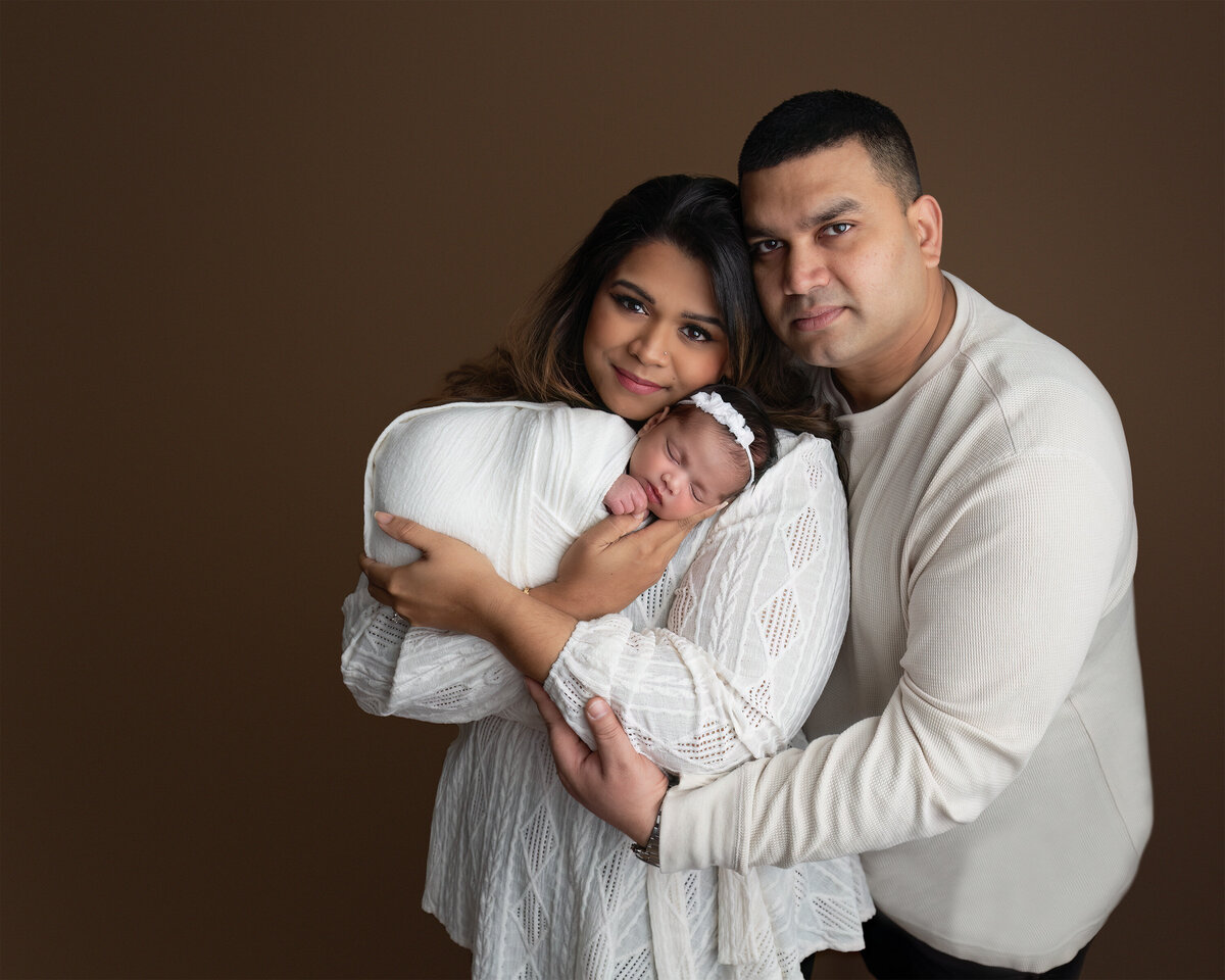 A classic family studio portrait featuring parents holding their newborn in front of a seamless mocha background. Captured at Meraki Photographic Creations in Paris, Ontario.