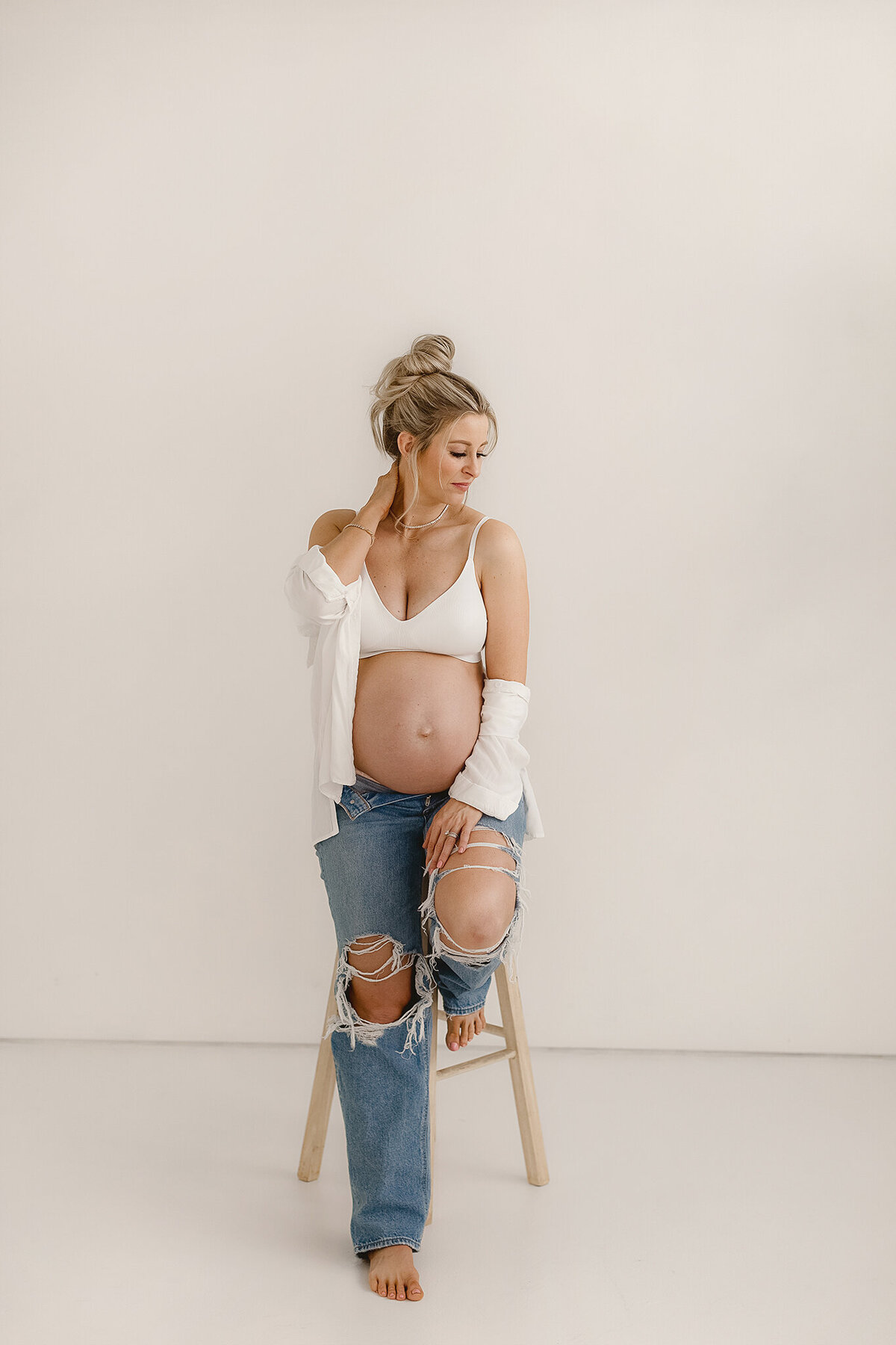 mom sitting on stool in modern denver photography studio holding her hair and bump for some intimate photos pregnant