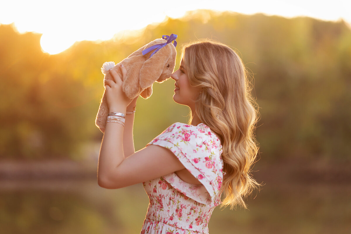 Dallas senior photographer captures a young woman in a floral dress, gently holding a stuffed bunny to her face during golden hour by a river.