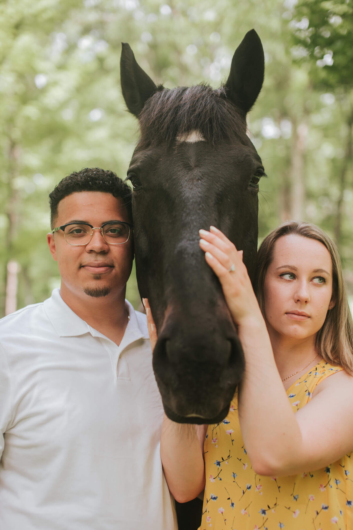 A couple poses with a black horse during their Hudson Valley engagement session, with the woman gently touching the horse’s face while showcasing her engagement ring.