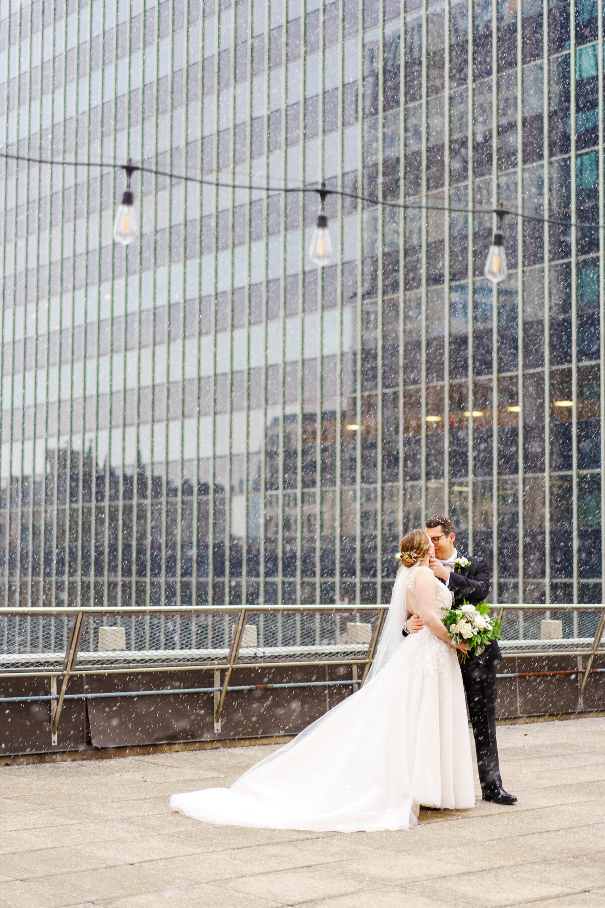 Bride and groom kiss under industrial lights atop a roof in downtown Columbus, Ohio as the snow falls.