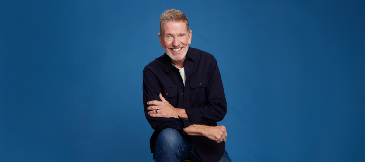 Michael Hyatt seated against a solid blue background, smiling and casually dressed in a dark shirt and jeans.