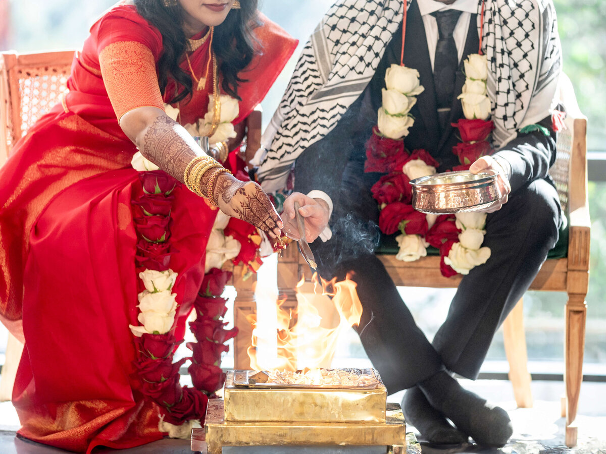 A couple in formal attire performs a wedding ritual by a ceremonial fire. The woman, in a red sari and gold jewelry, is holding an offering. Both are adorned with garlands, and the man wears a patterned shawl.